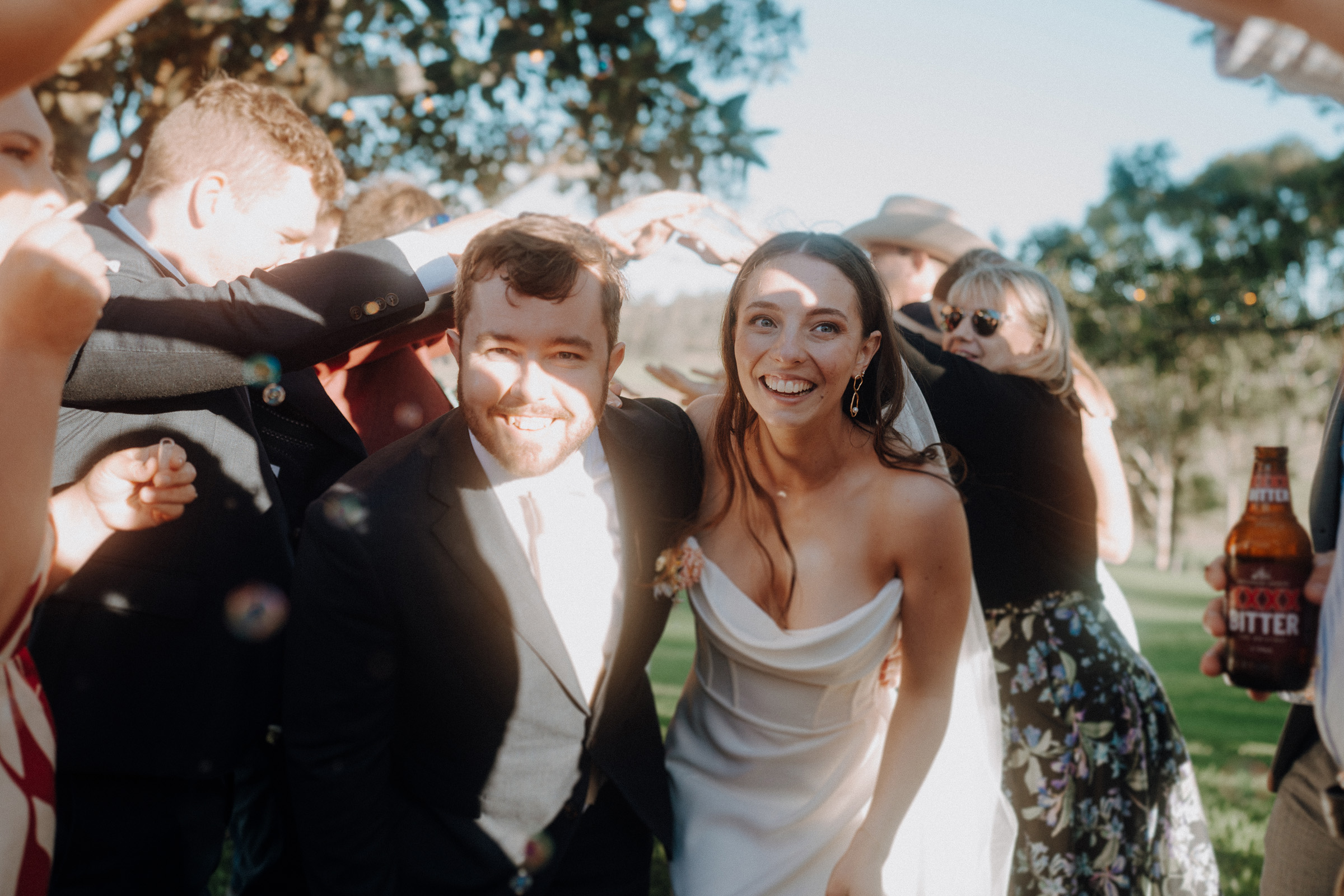 Bride and groom smiling and walking through a tunnel made by guests' arms at an outdoor wedding celebration.