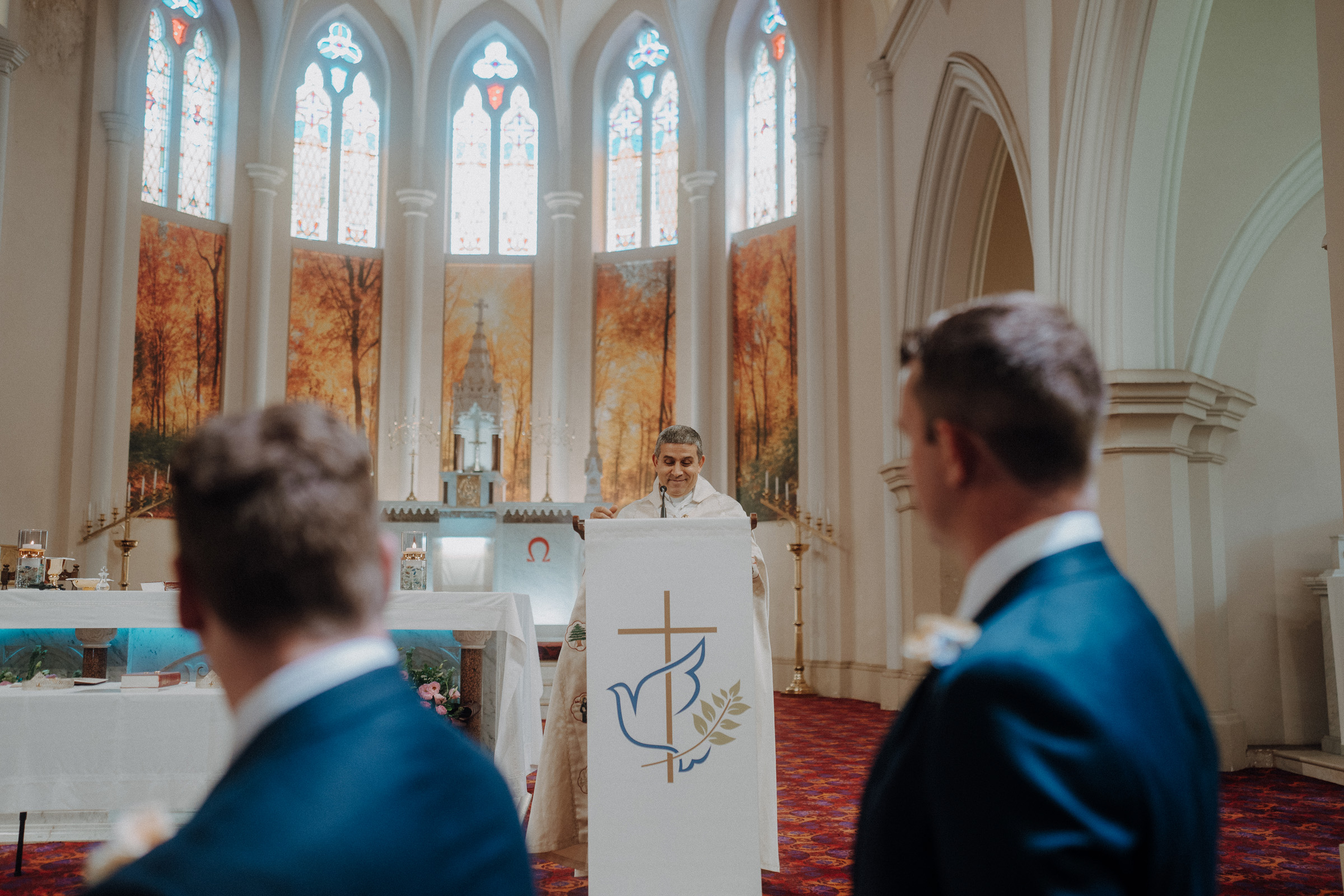 A priest stands at a lectern inside a church with stained glass windows, addressing two men in suits who are seated.