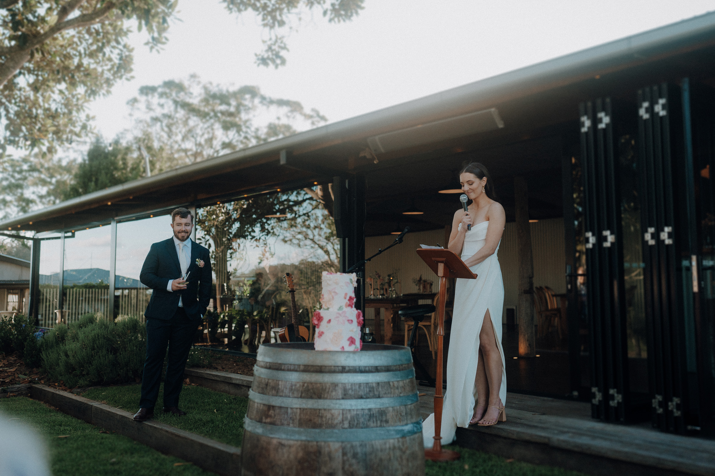 A bride in a white dress gives a speech at a microphone stand beside a gift table. A groom stands nearby holding a glass. They are outdoors near a building with open doors.