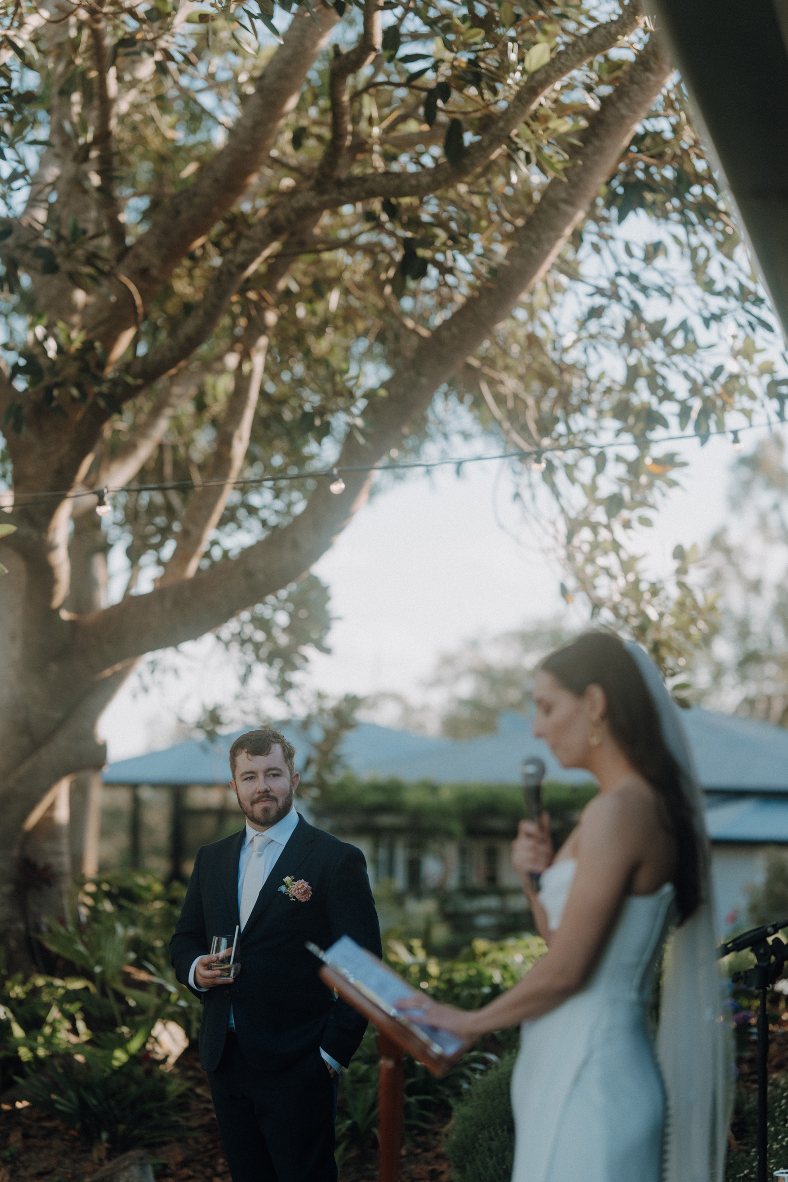 A couple stands outdoors during a wedding ceremony. The woman holds a microphone and reads from a paper, while the man in a suit listens, holding a glass. Trees and a building are in the background.