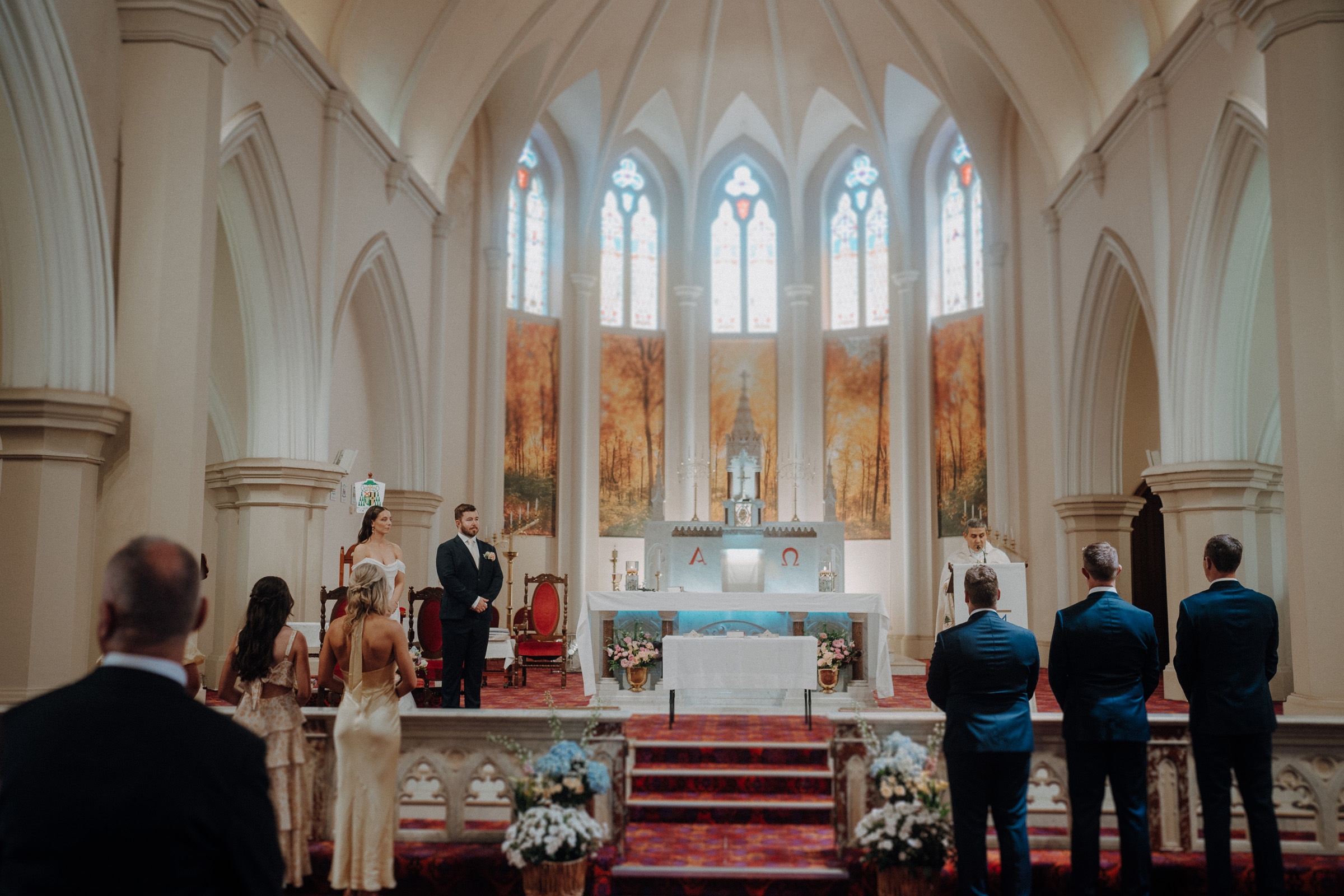 A wedding ceremony in a church with a bride, groom, and guests facing an altar. The interior features arched ceilings and stained glass windows.