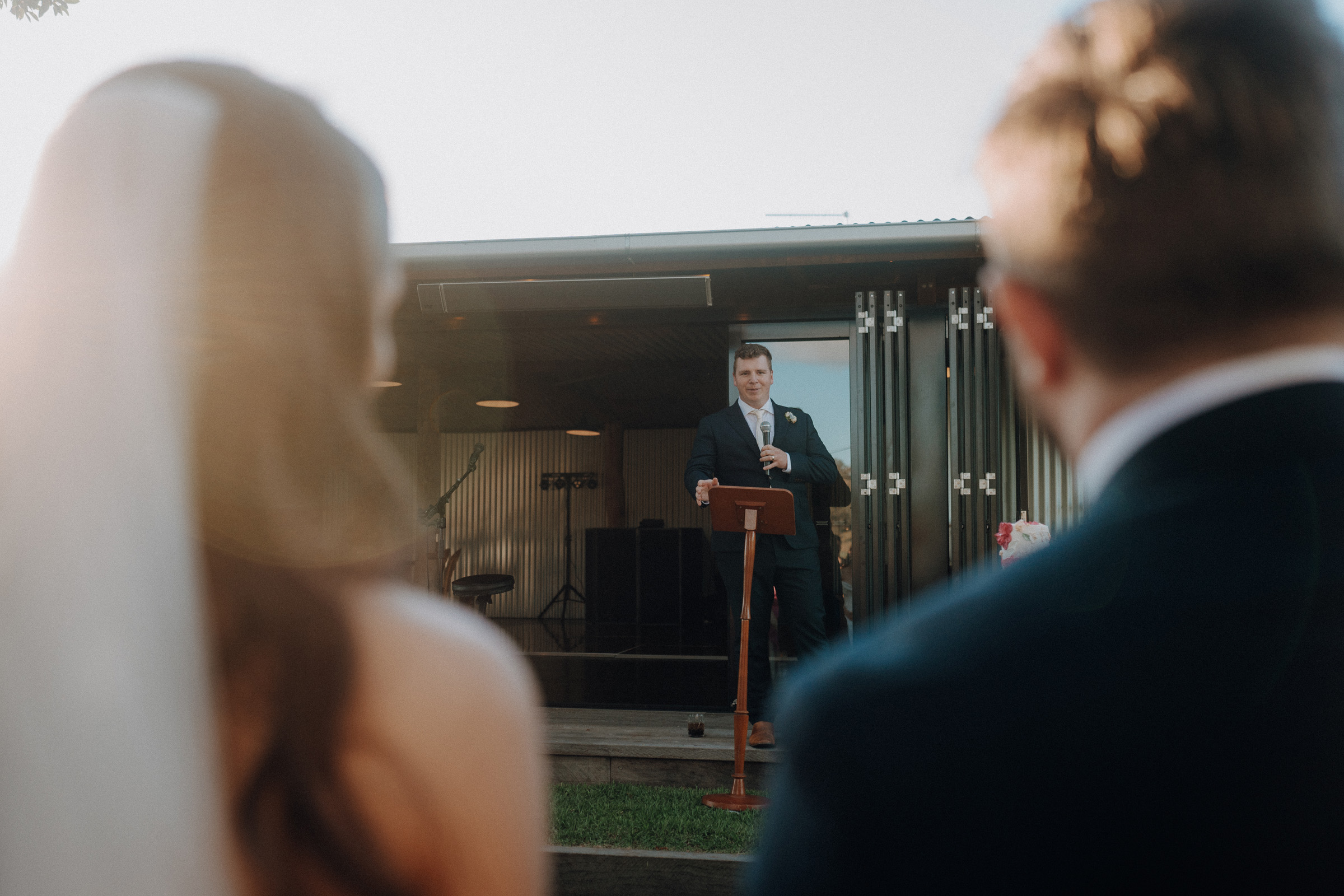 A man in a suit speaks at a podium outdoors, facing an audience. A couple, partially visible, listens in the foreground.