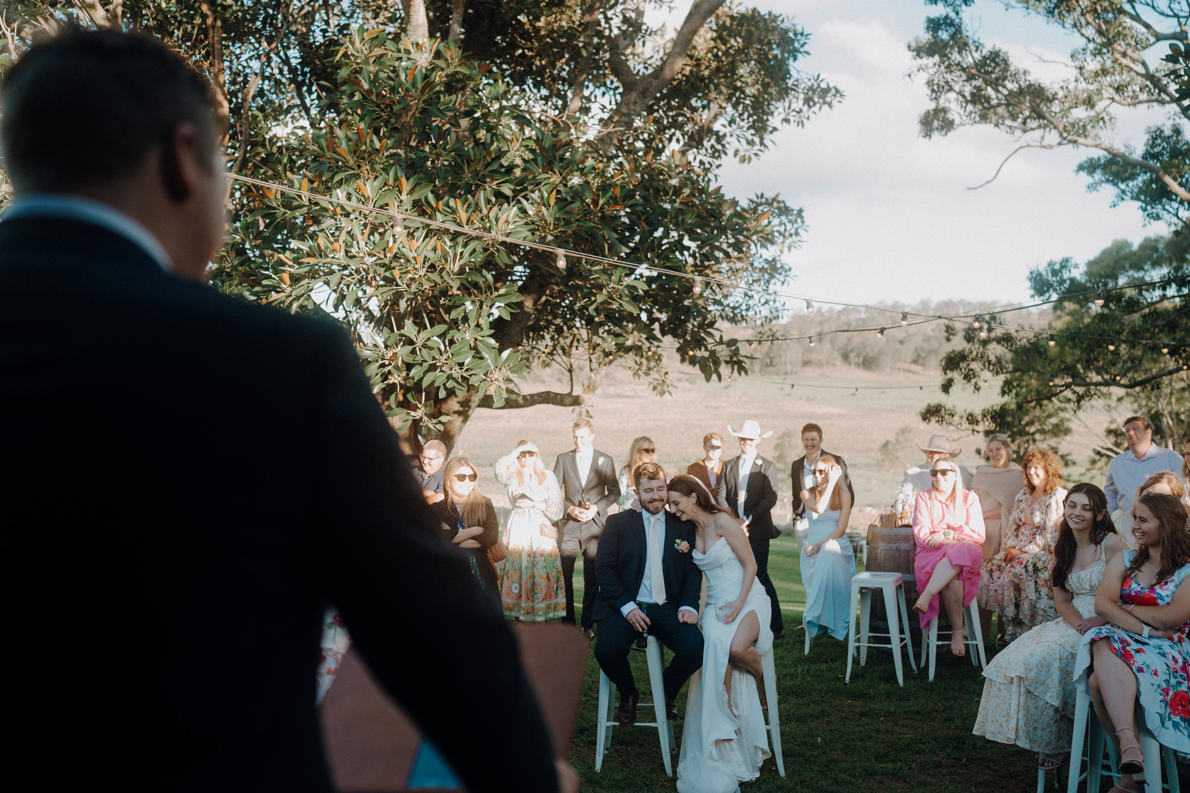 A wedding ceremony outdoors with guests seated under trees. A man speaks at a podium while a couple sits on stools in front of the audience.