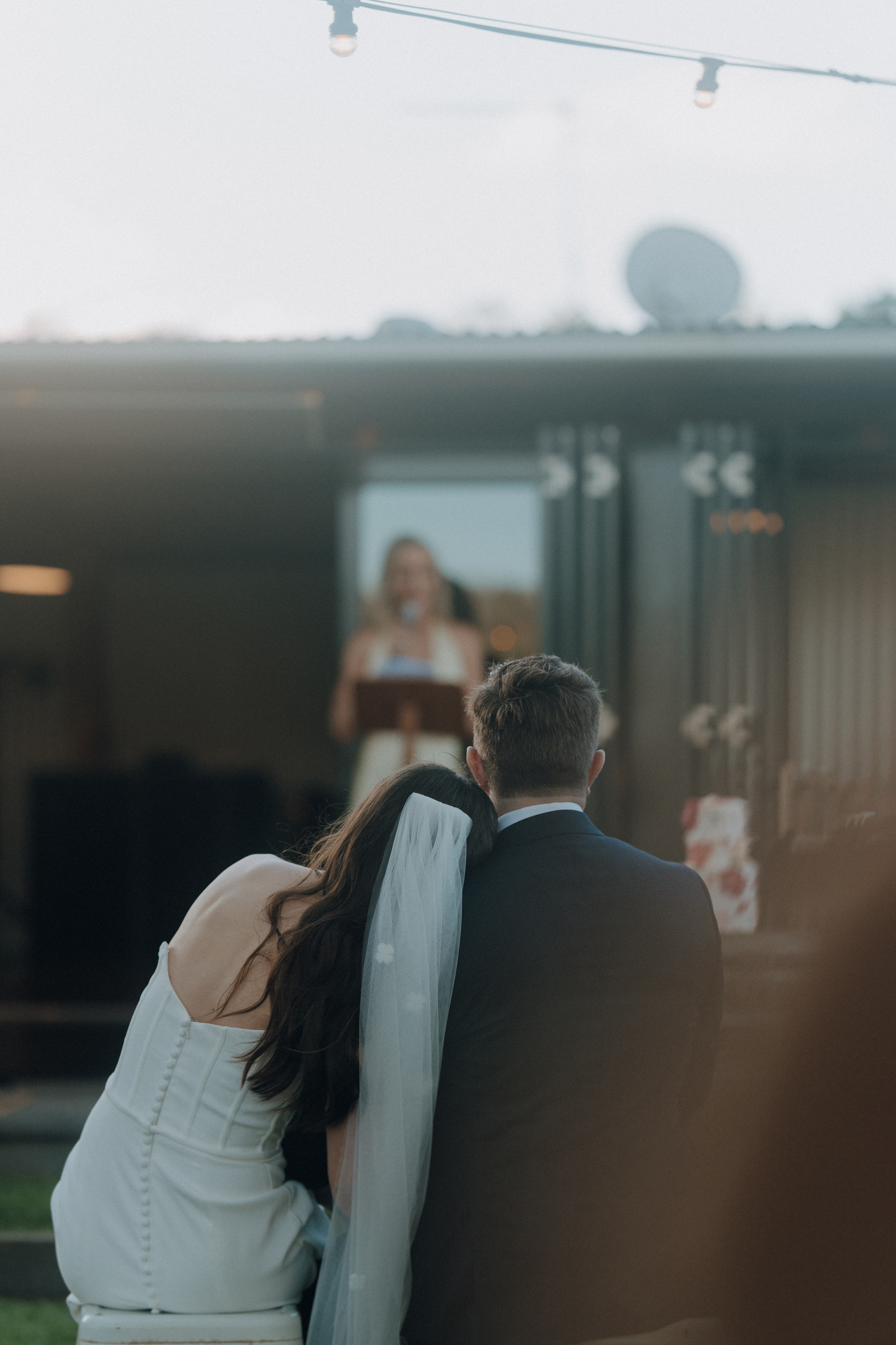 A couple dressed in wedding attire sits closely together, watching a woman speak at a podium in an outdoor setting.