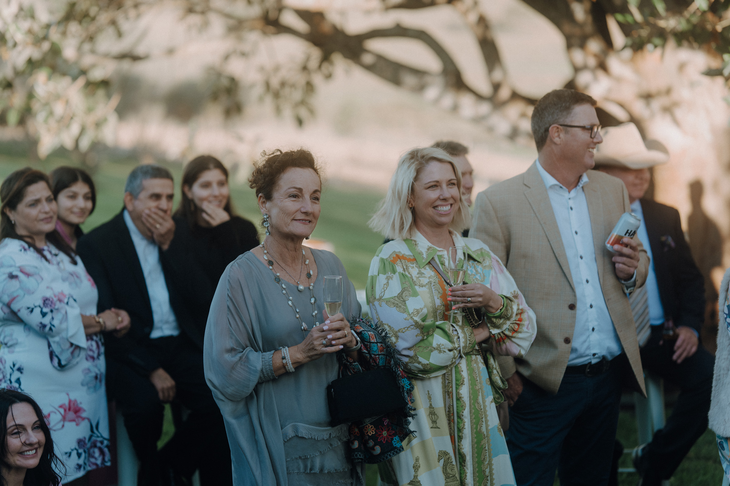 A group of people standing outdoors in a relaxed setting, some holding drinks, with trees and grass in the background.