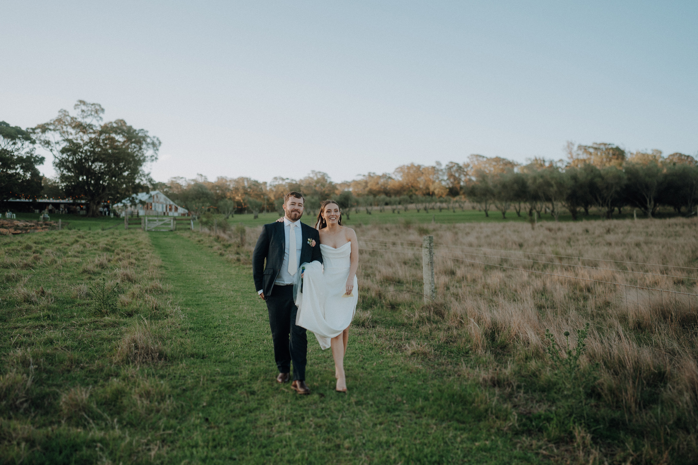 A bride and groom walk arm in arm through a grassy field with trees in the background.