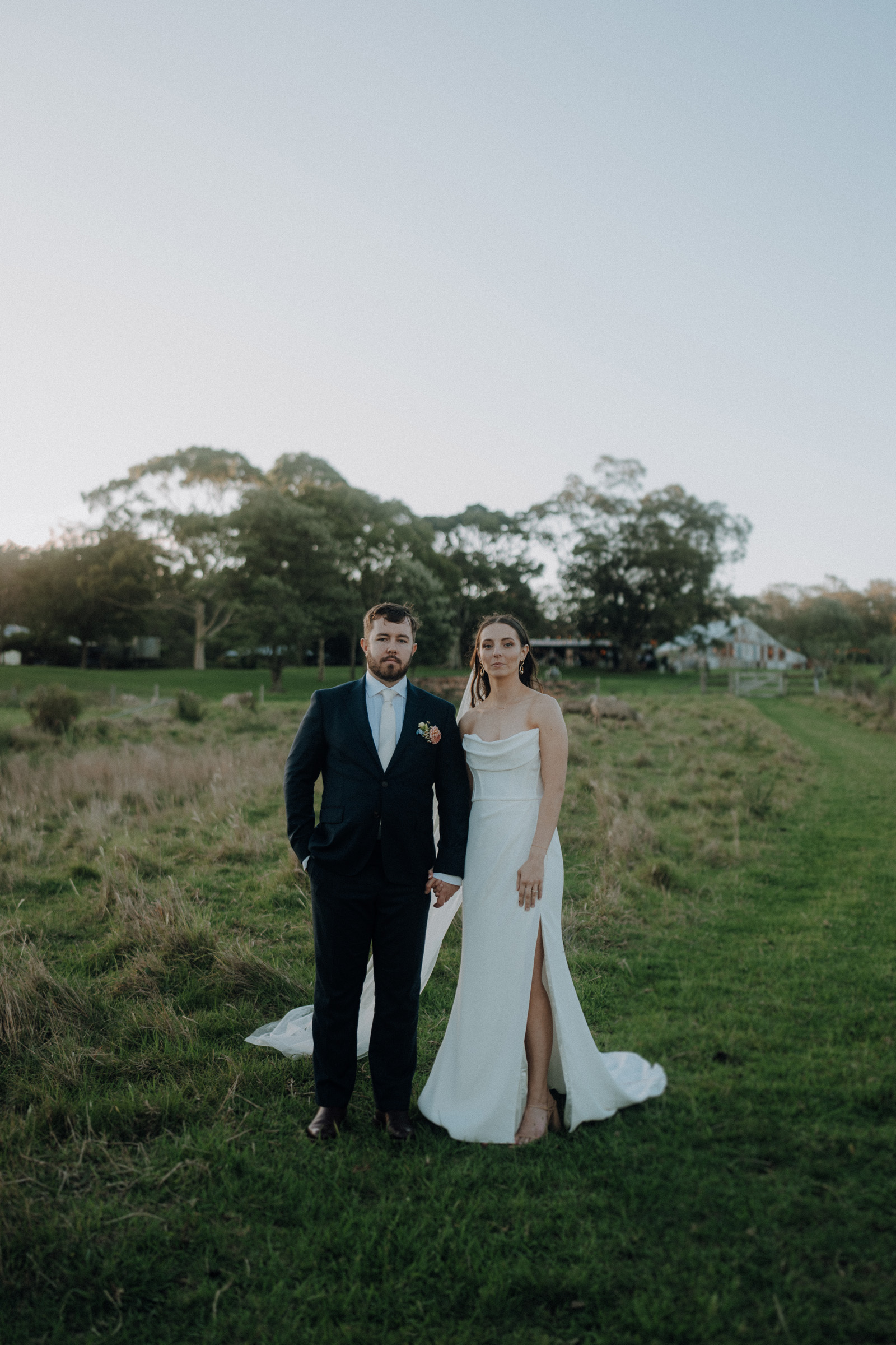 A bride in a white dress and a groom in a dark suit stand side by side on a grassy field with trees in the background.