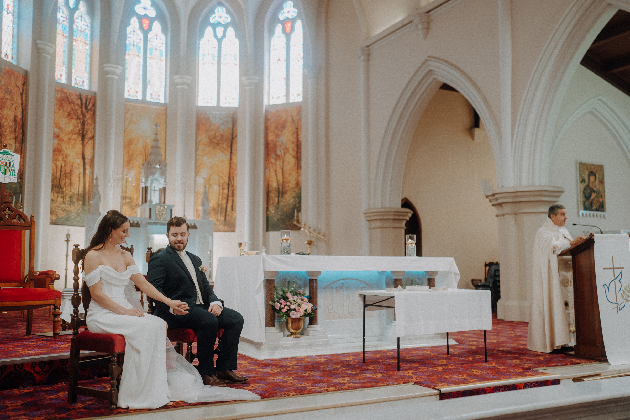 A bride and groom sit holding hands in a church with stained glass windows, while a priest stands at a podium on the right.