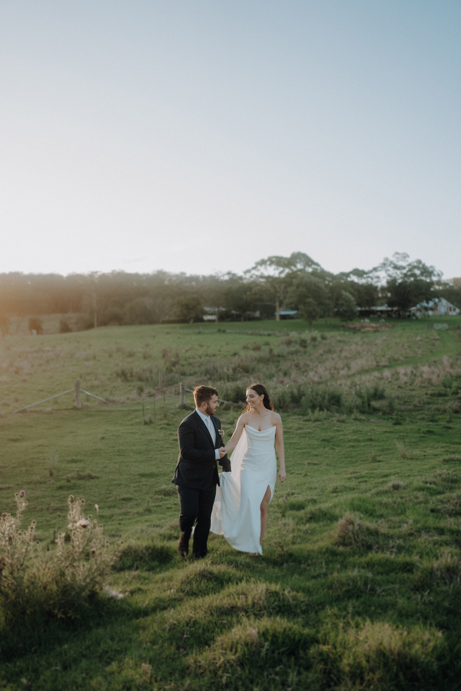 A couple in formal attire walks hand in hand across a grassy field. The woman wears a white dress, and the man is in a dark suit. Trees and a fence are visible in the background.