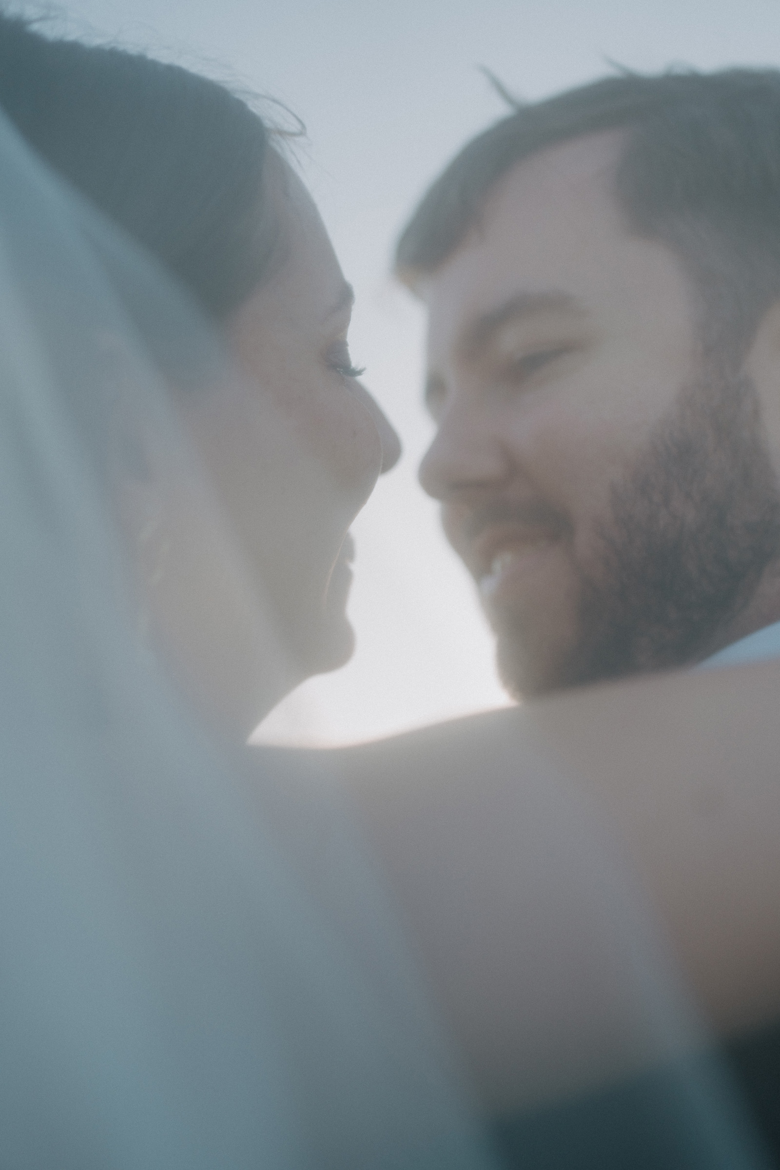 Close-up of a couple smiling at each other on their wedding day, with the woman wearing a veil. The image is softly focused and slightly blurred, creating a dreamy effect.