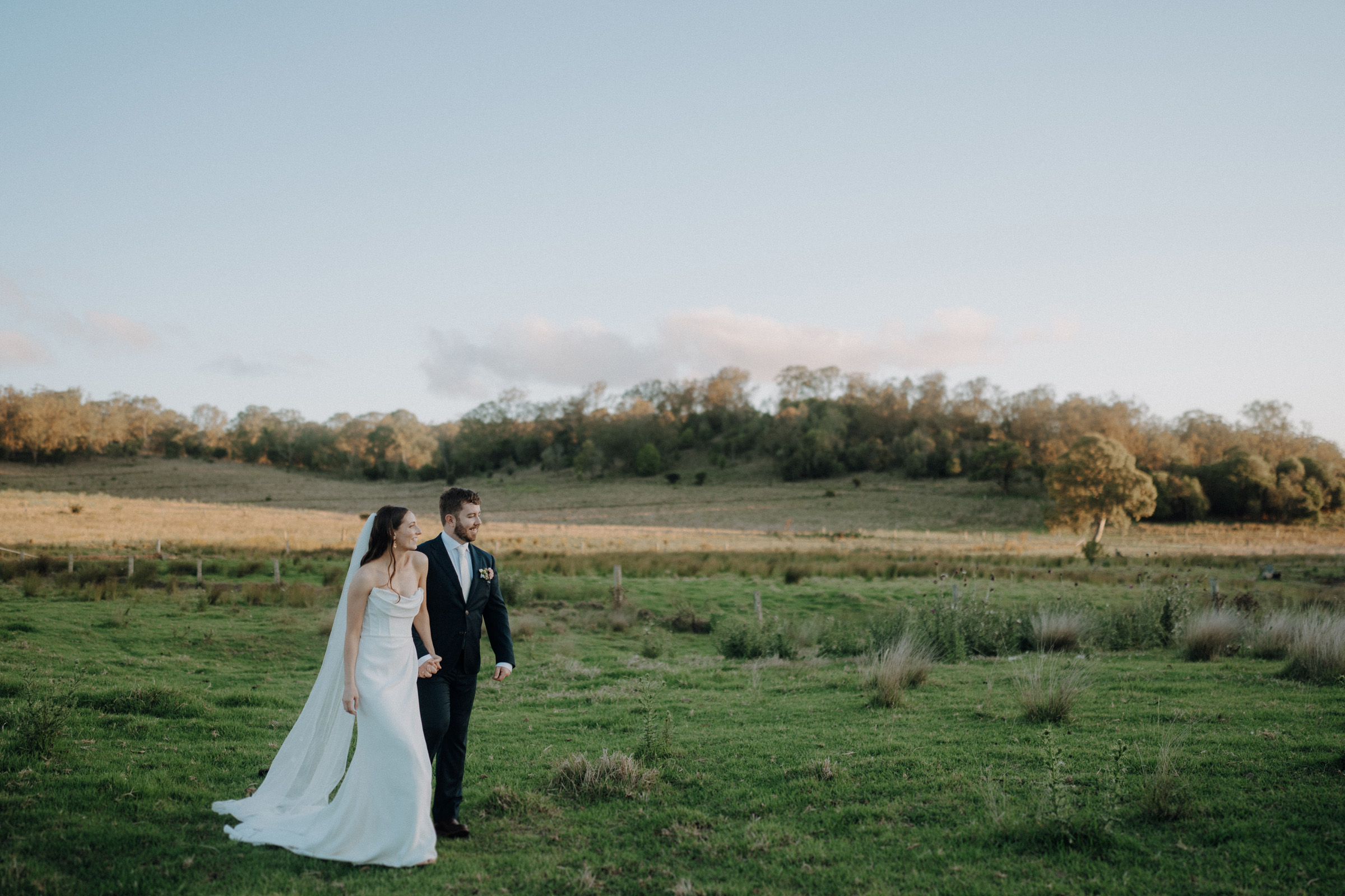 A couple in wedding attire walks hand in hand across a grassy field, with a backdrop of trees under a clear sky.