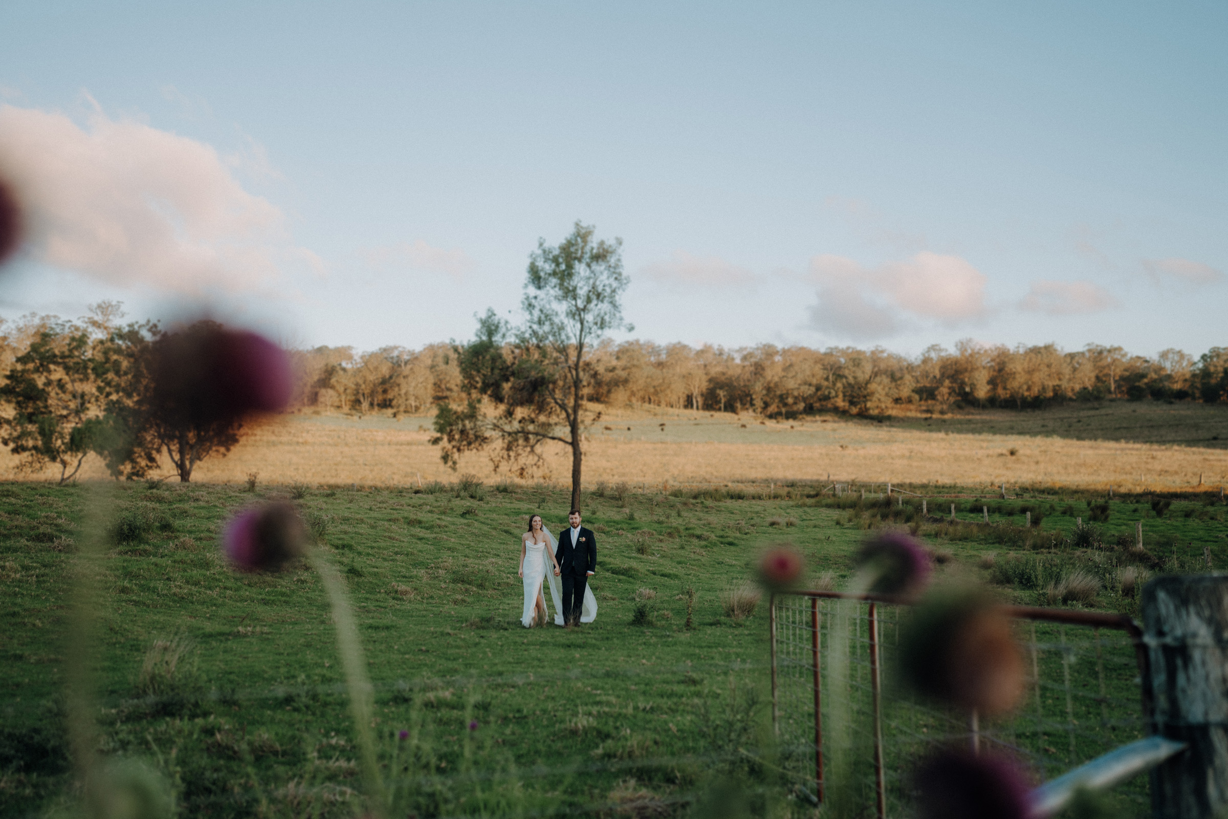 A bride and groom walk across a grassy field, with trees and a clear sky in the background. Blurred flowers are in the foreground.