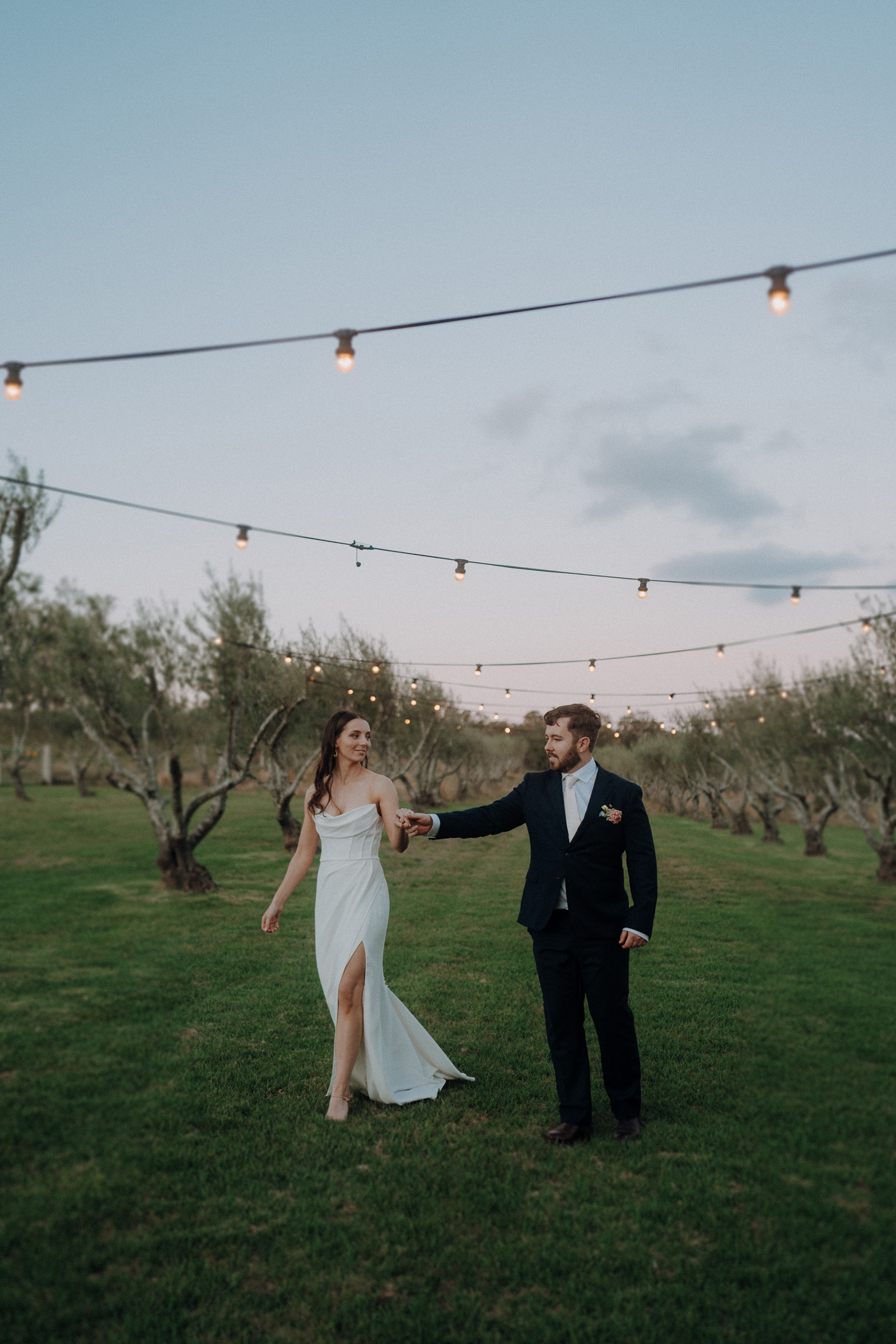 A couple in formal attire walks hand in hand through a grassy field decorated with string lights.