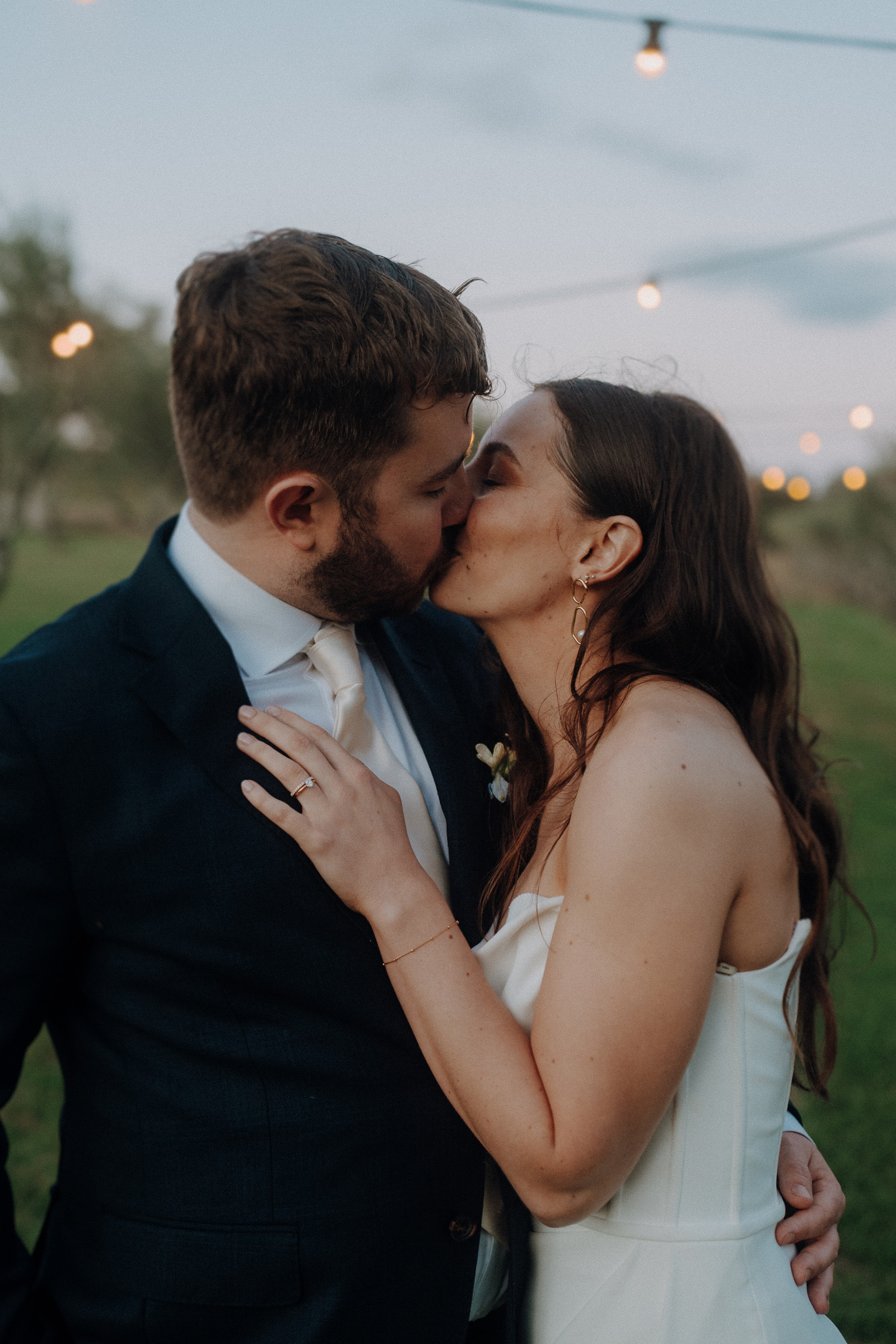 A couple dressed in formal attire shares a kiss outdoors at dusk, with string lights softly glowing in the background.