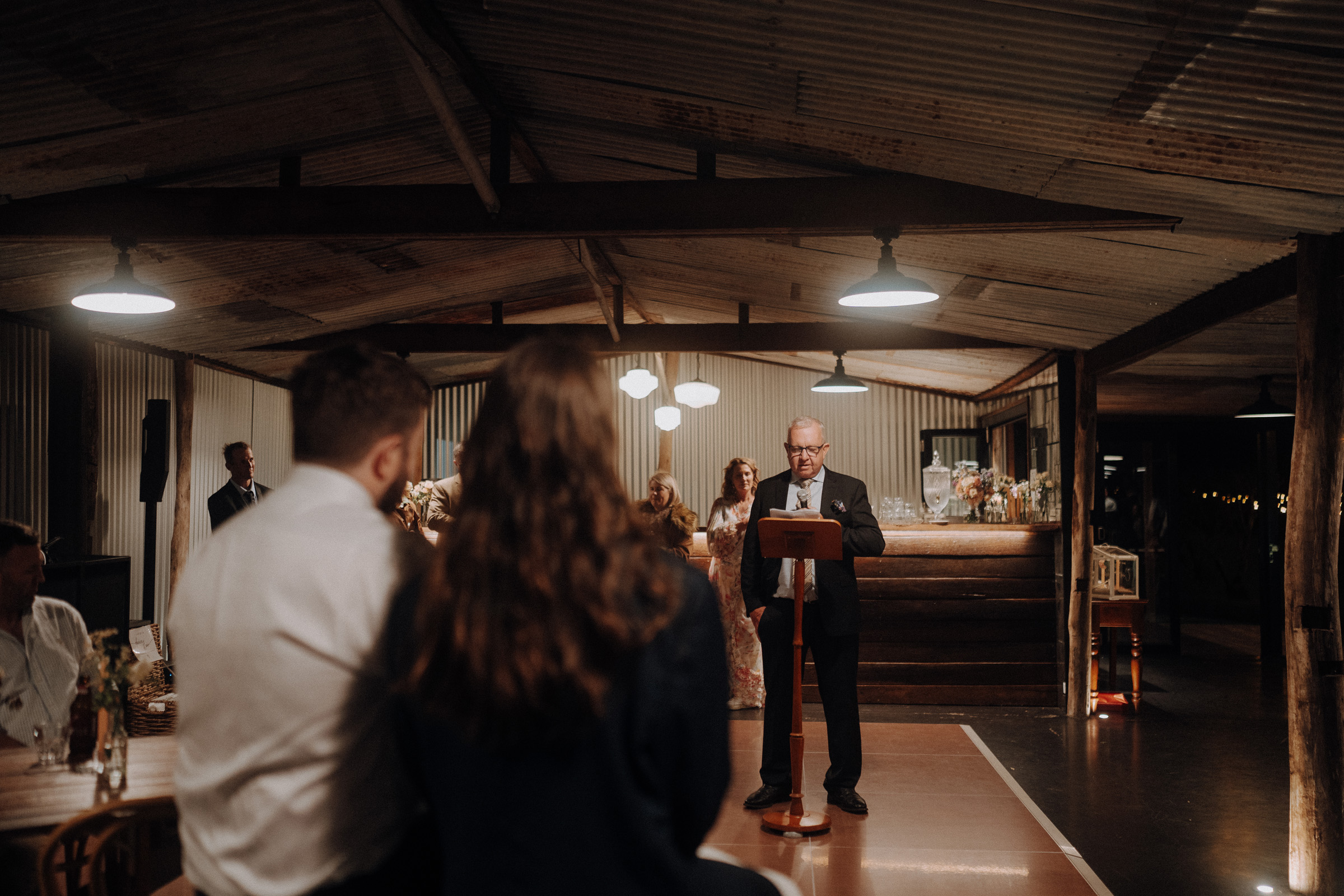 A person in a suit speaks at a podium in a dimly lit rustic venue. Audience members are seated in the foreground, with wooden beams and decor visible in the background.