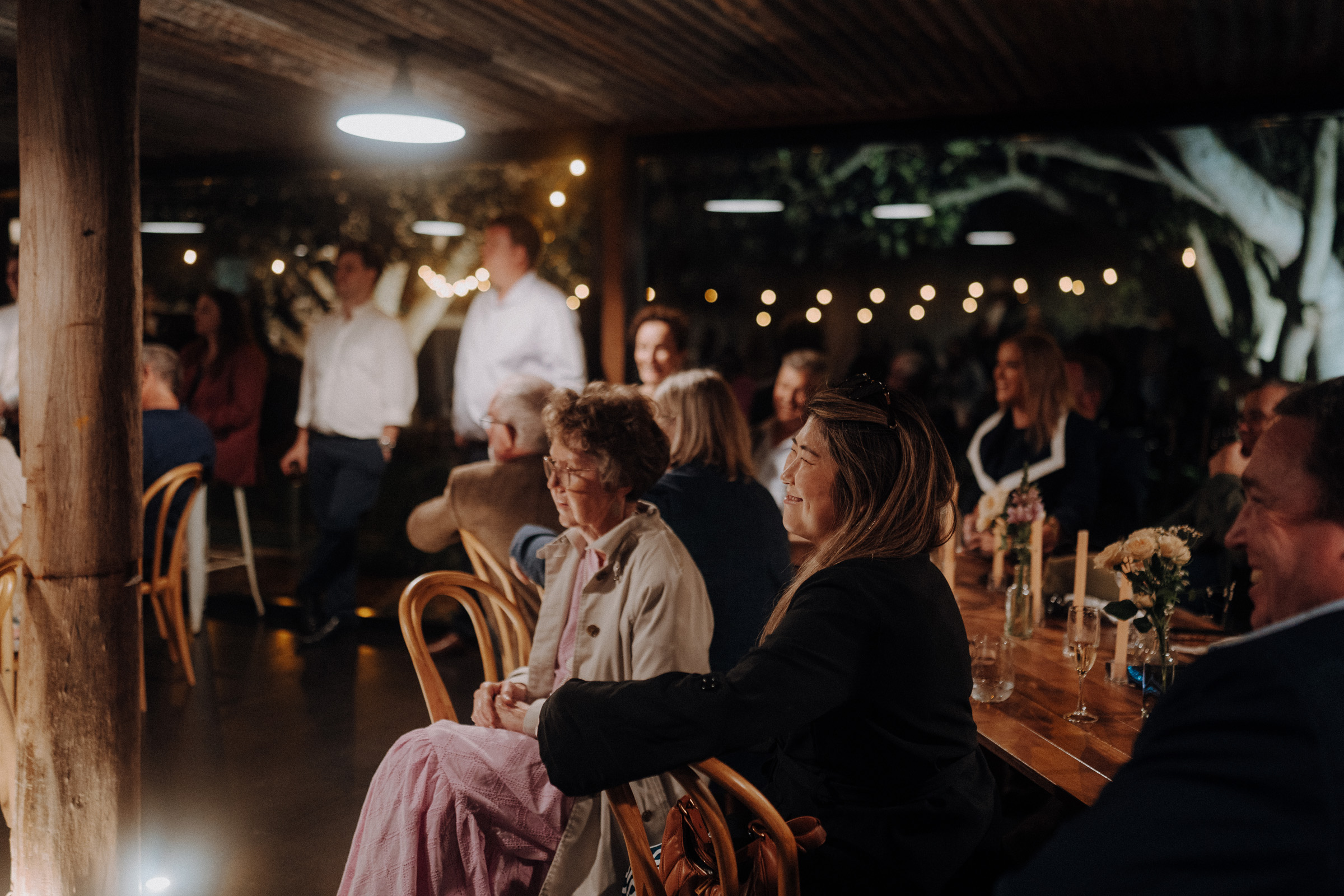 People seated and standing under a covered outdoor area with string lights, attentively watching an unseen event. Tables are decorated with flowers and candles.