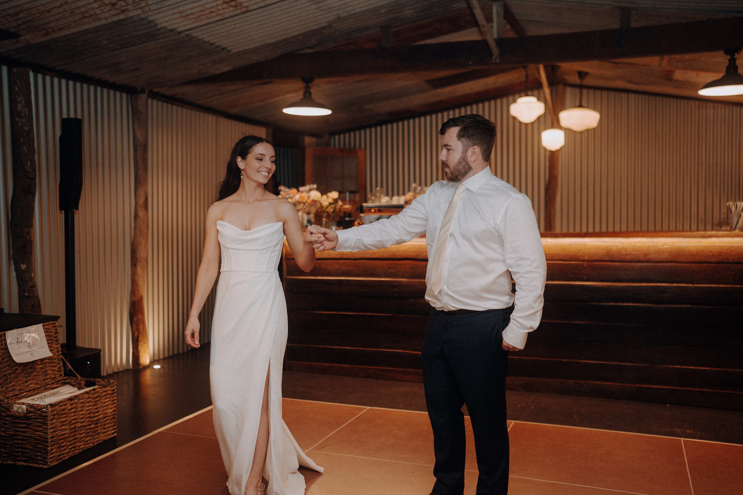 A couple dances in a rustic setting. The woman wears a white dress with a slit, and the man wears a white shirt and dark pants. They stand on a square wooden floor.