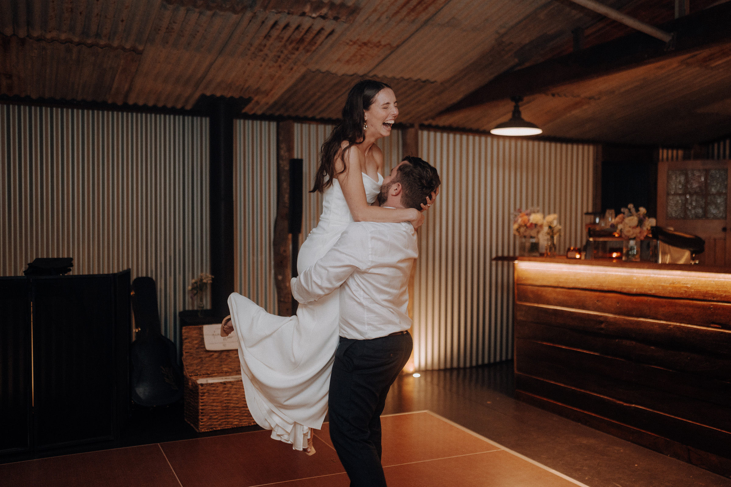 A bride in a white dress is lifted by a groom in a white shirt on a dance floor in a rustic venue.