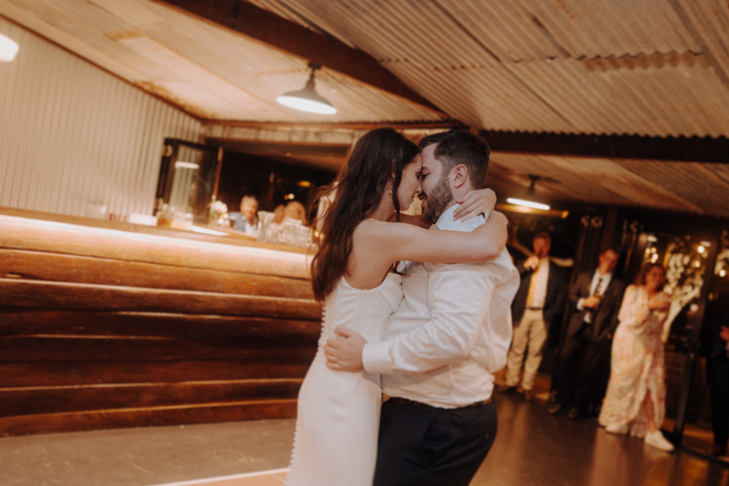 A couple embraces while dancing at a rustic indoor event. People in the background watch.