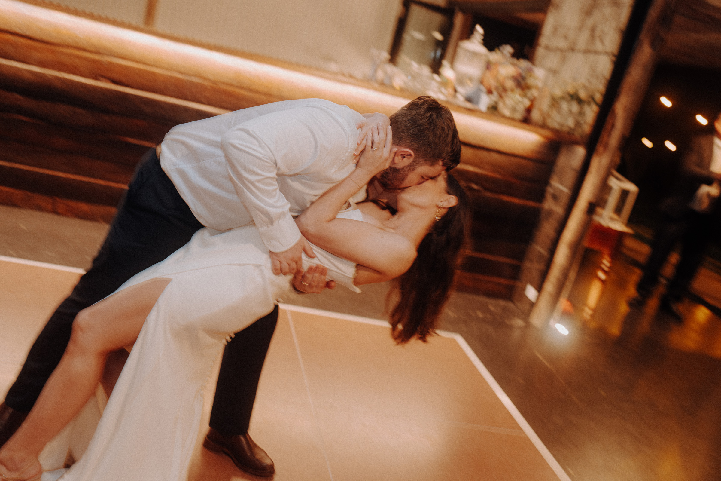 A couple in formal attire shares a kiss while performing a dip dance move on a wooden floor.