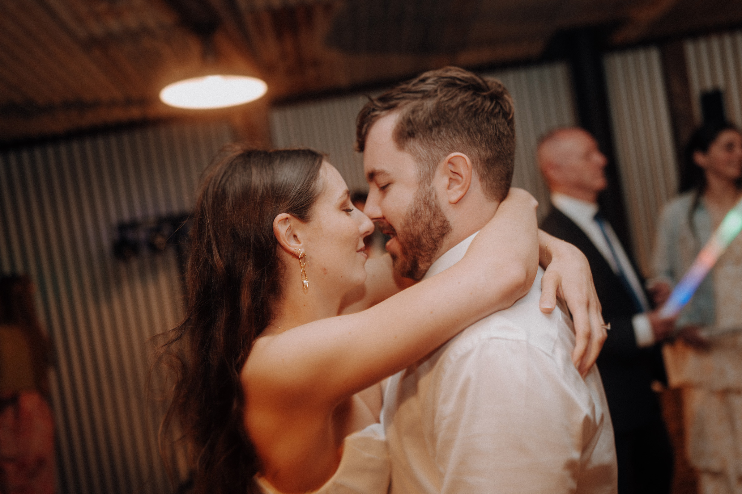 A couple embraces closely, smiling with closed eyes at a social gathering. People in formal attire are in the background.