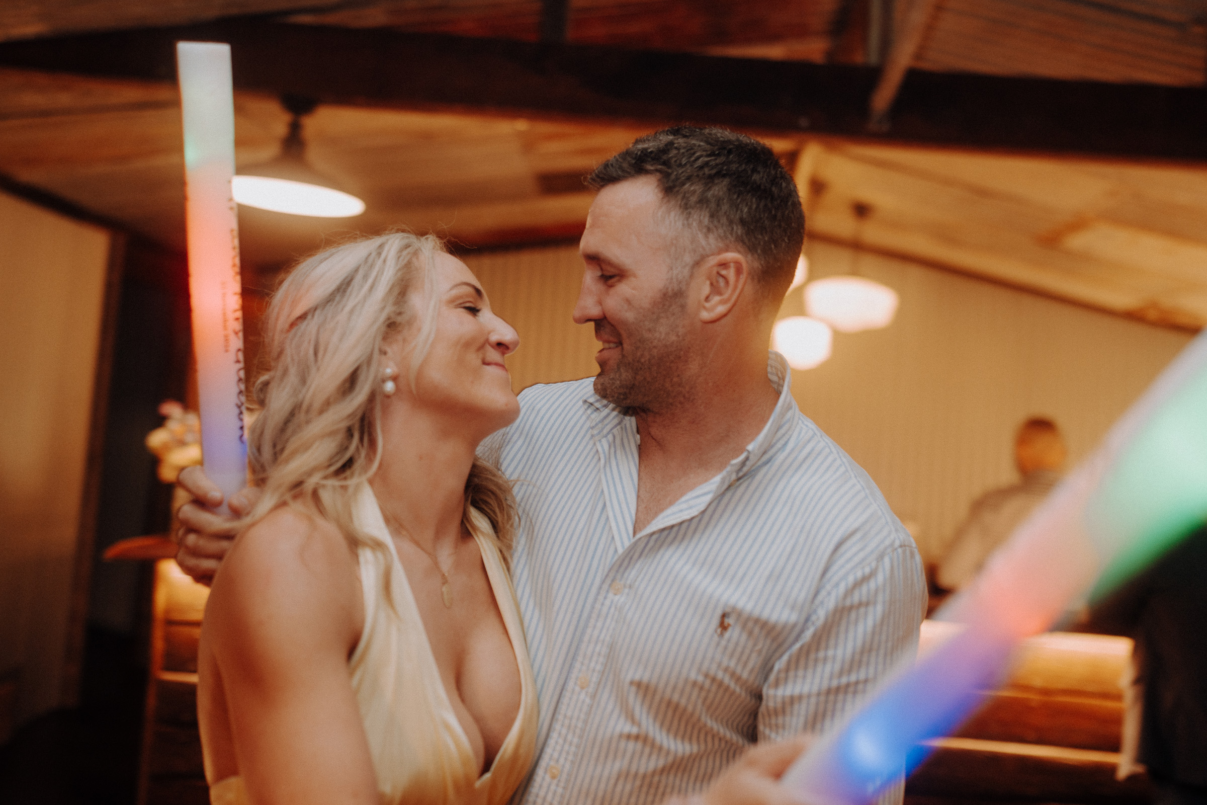 A man and woman smiling at each other in a warmly lit indoor setting, holding colorful light sticks.