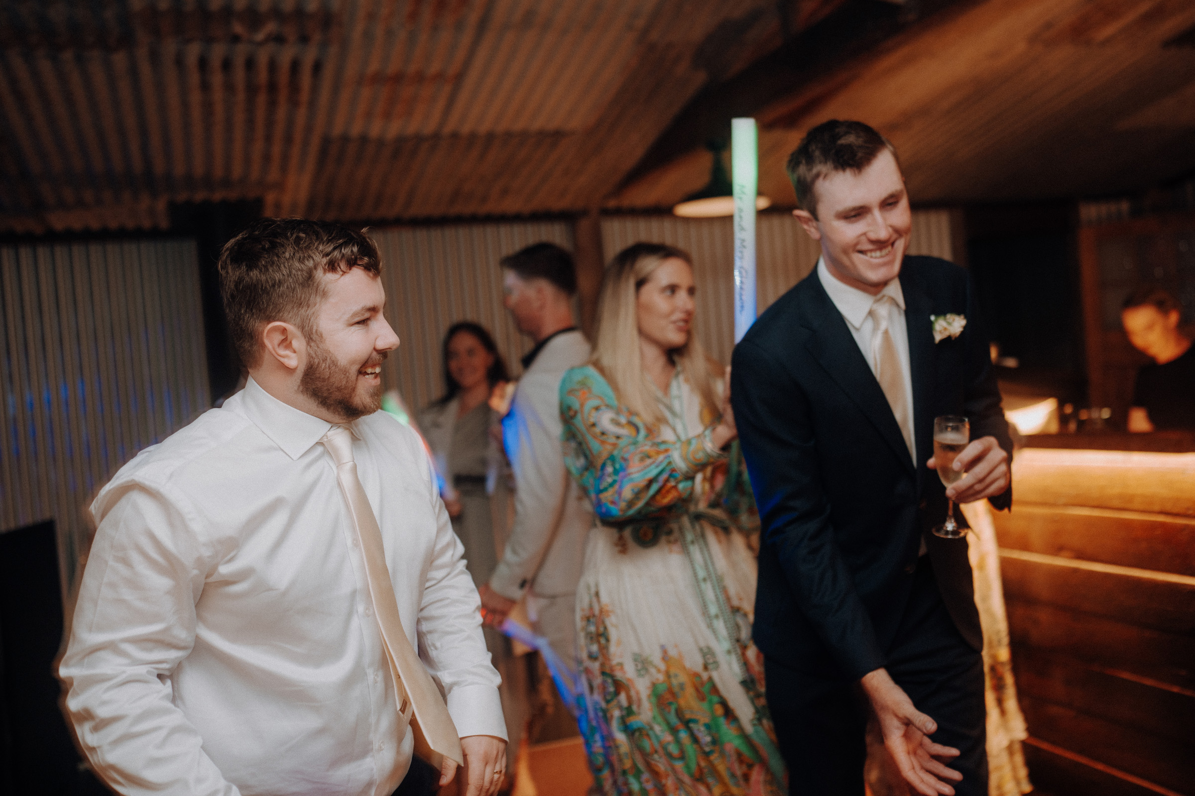 Two men in suits and ties are smiling and in conversation at a social event. One holds a champagne glass. Other people are in the background in a rustic indoor setting.