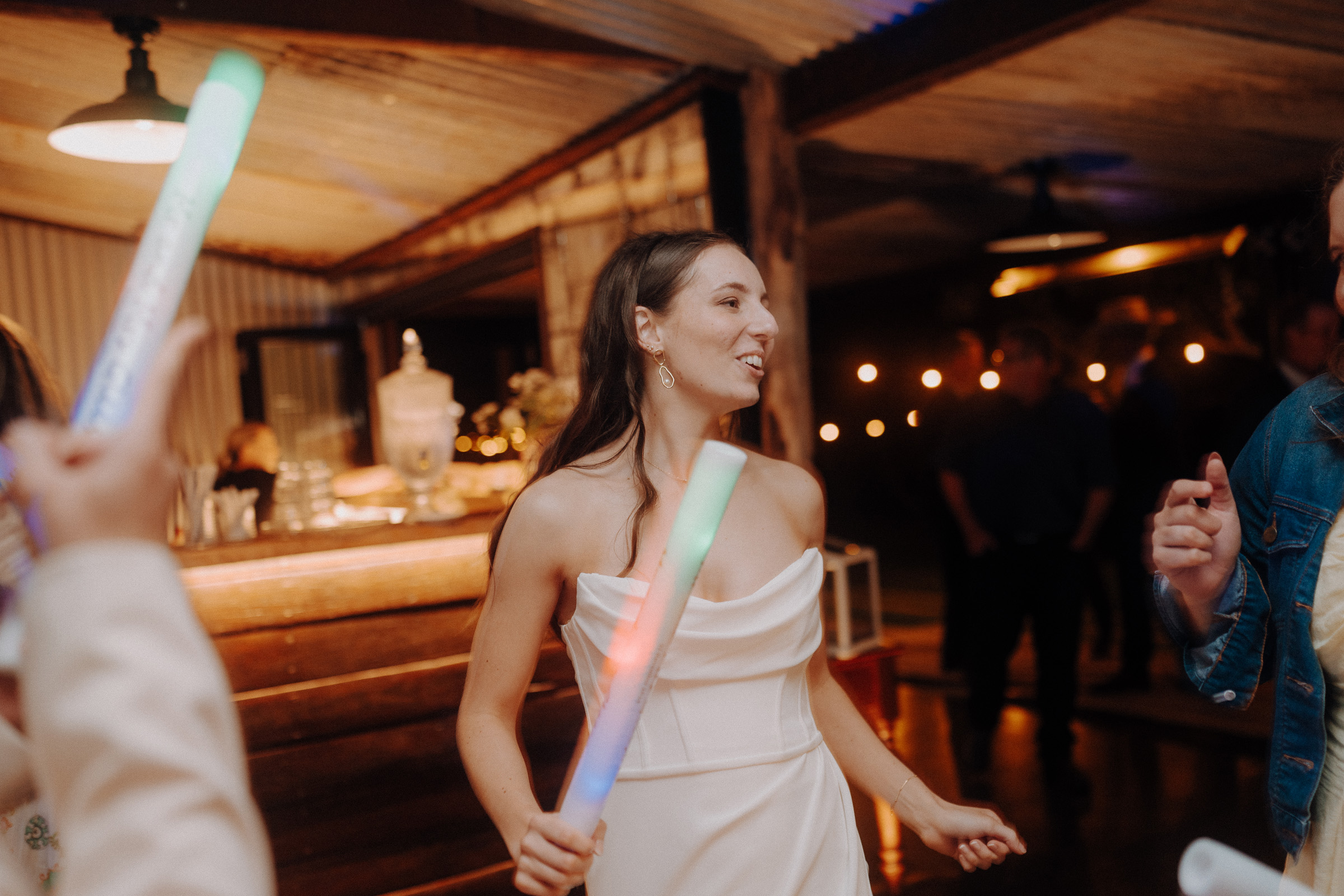 A woman in a white dress holds glowing sticks at a dimly lit indoor event. Others are partially visible around her.