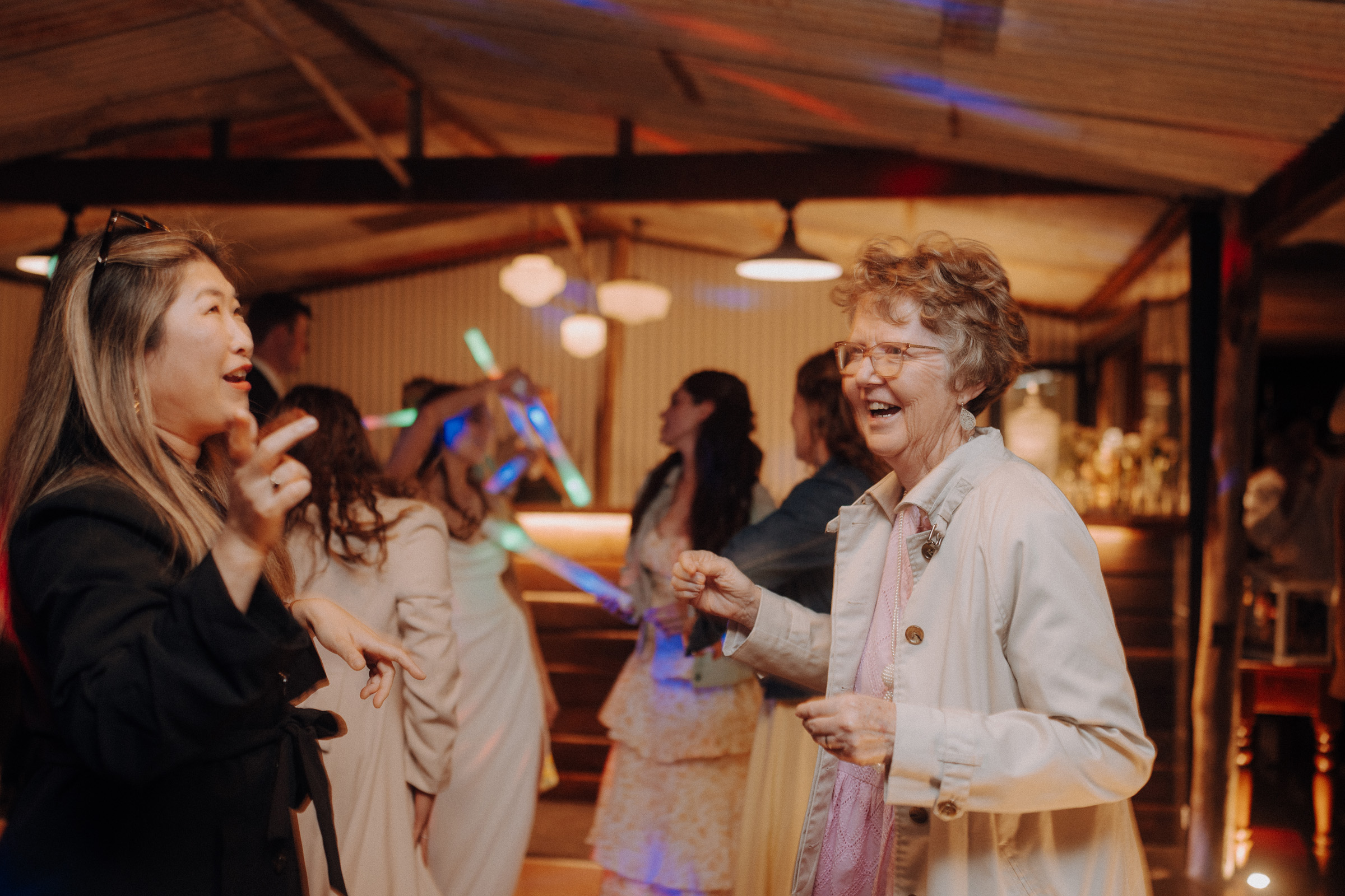 Two women laughing and dancing at a lively indoor gathering, surrounded by other people enjoying the event.