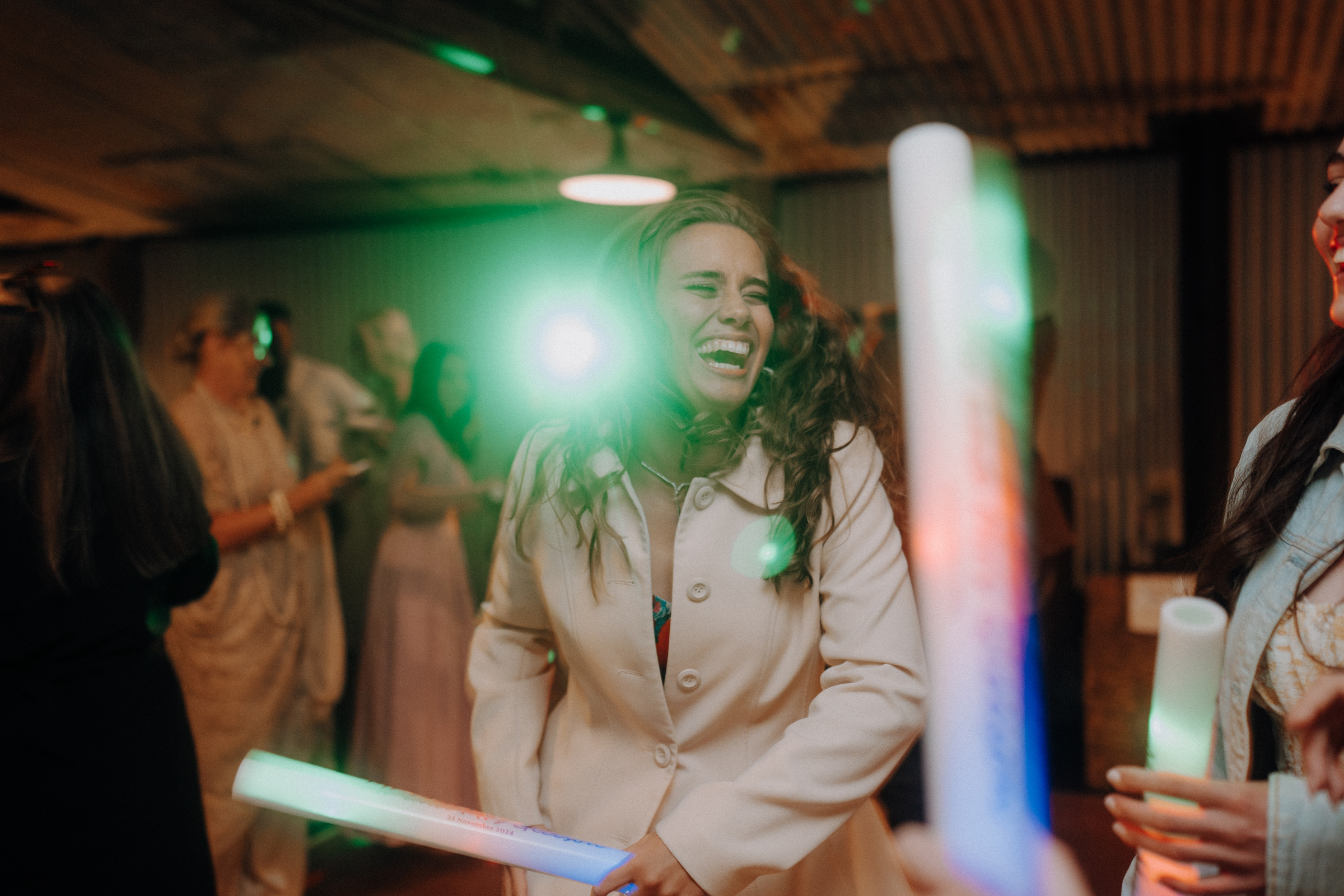 A woman in a light coat laughs joyfully at an indoor event with other people. She is surrounded by colorful lights and appears to be holding a glowing stick.