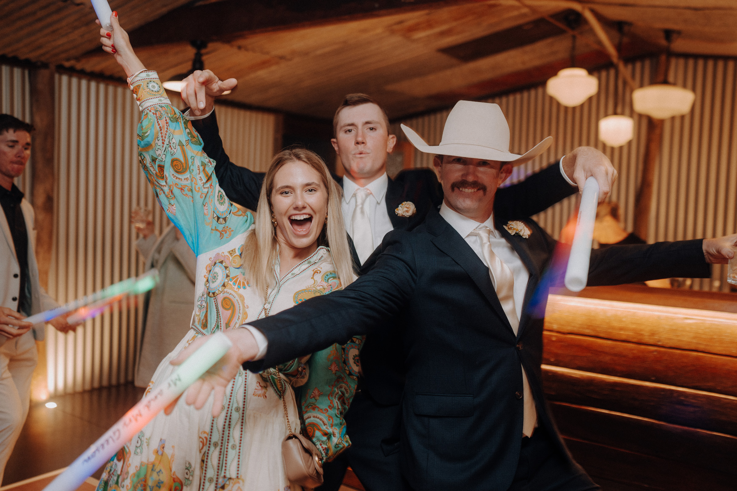 People in lively attire are celebrating indoors, holding glowing sticks. One person is wearing a cowboy hat. The room has wooden walls and hanging lights.