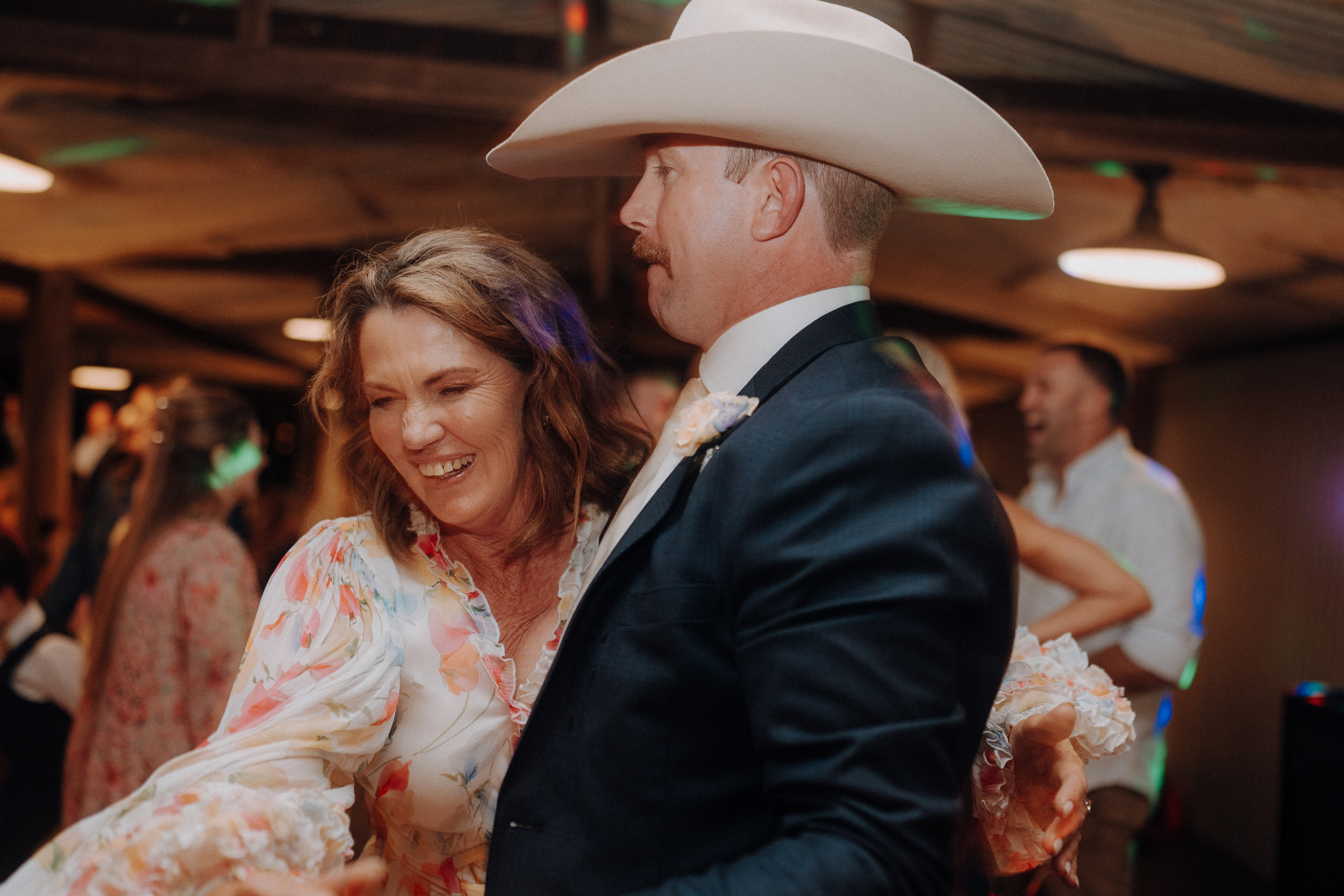 A man in a cowboy hat and a woman in a floral dress are dancing together, smiling, in a warmly lit room.