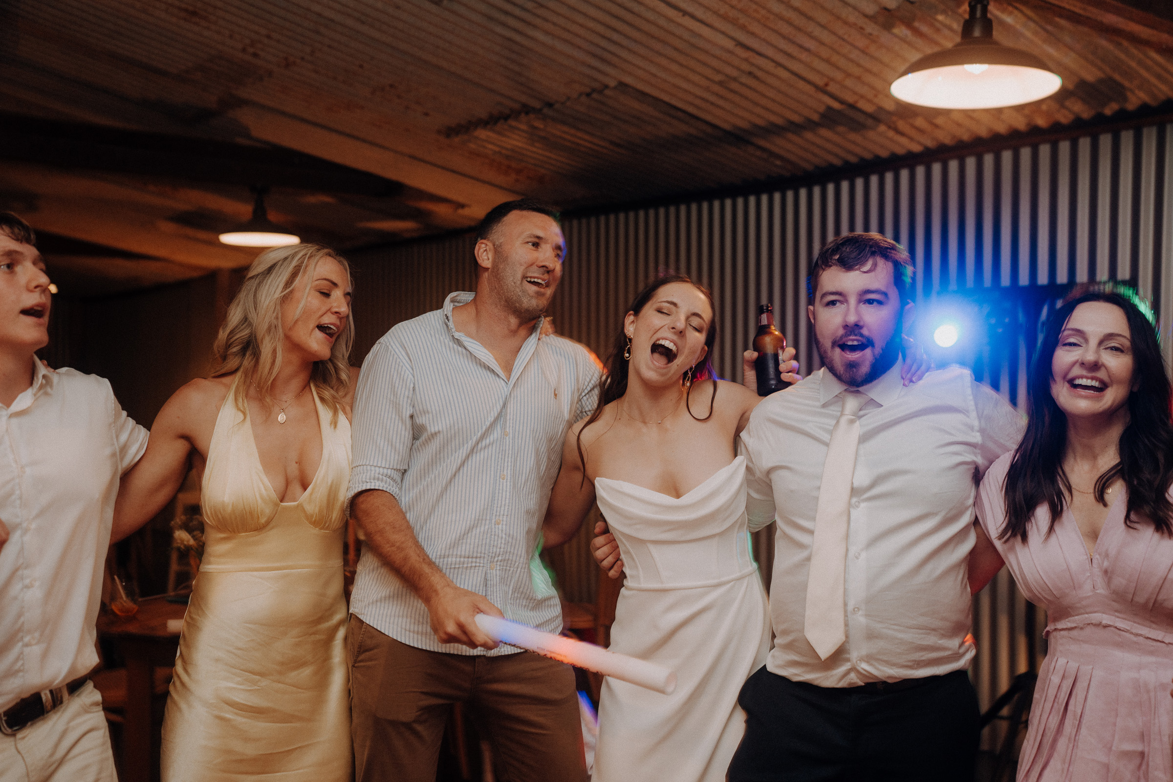 A group of six people in formal attire sing and embrace each other indoors, under warm lighting, holding drinks and a glow stick.