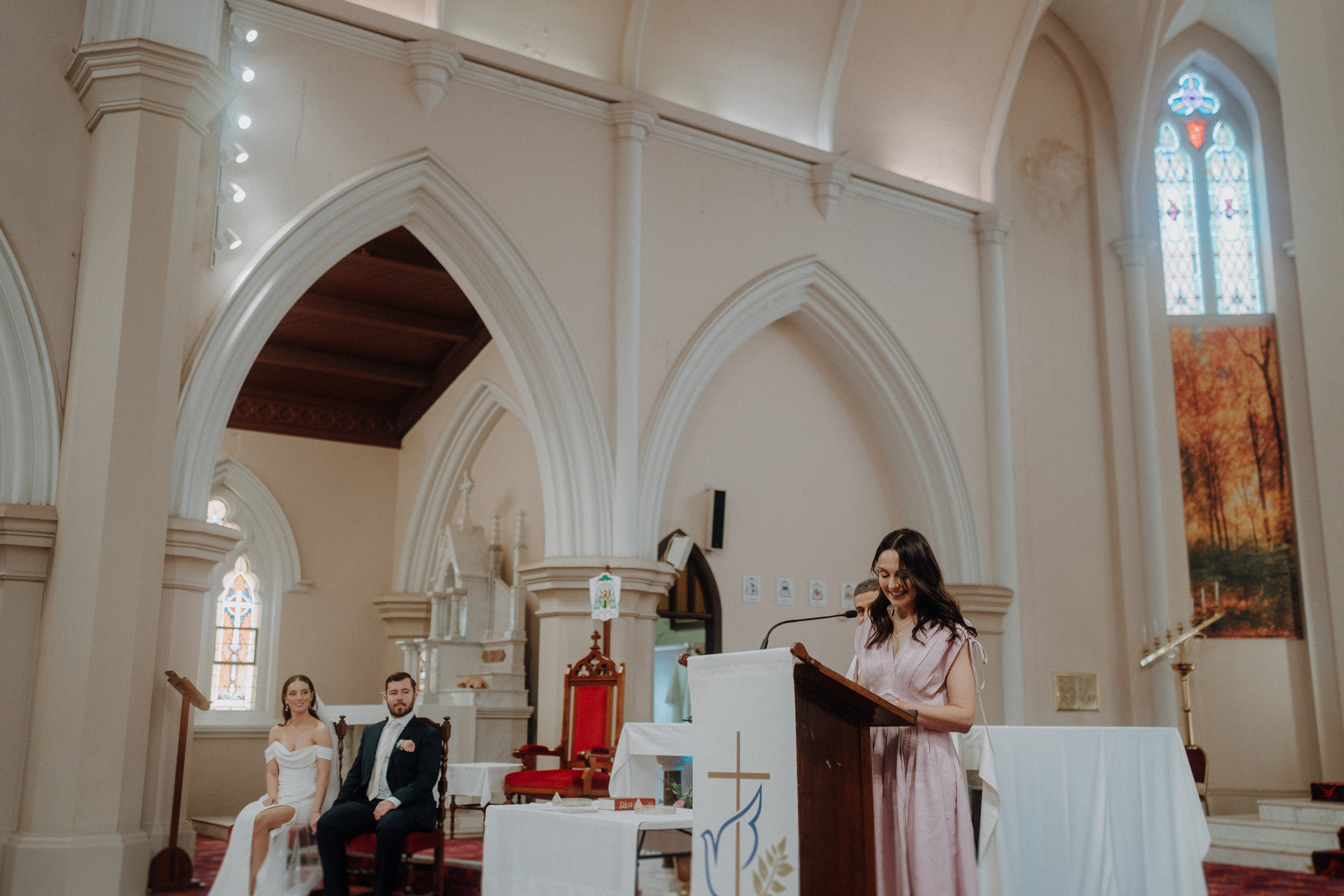 A woman speaks at a podium in a church, while a bride and groom sit on chairs in the background.