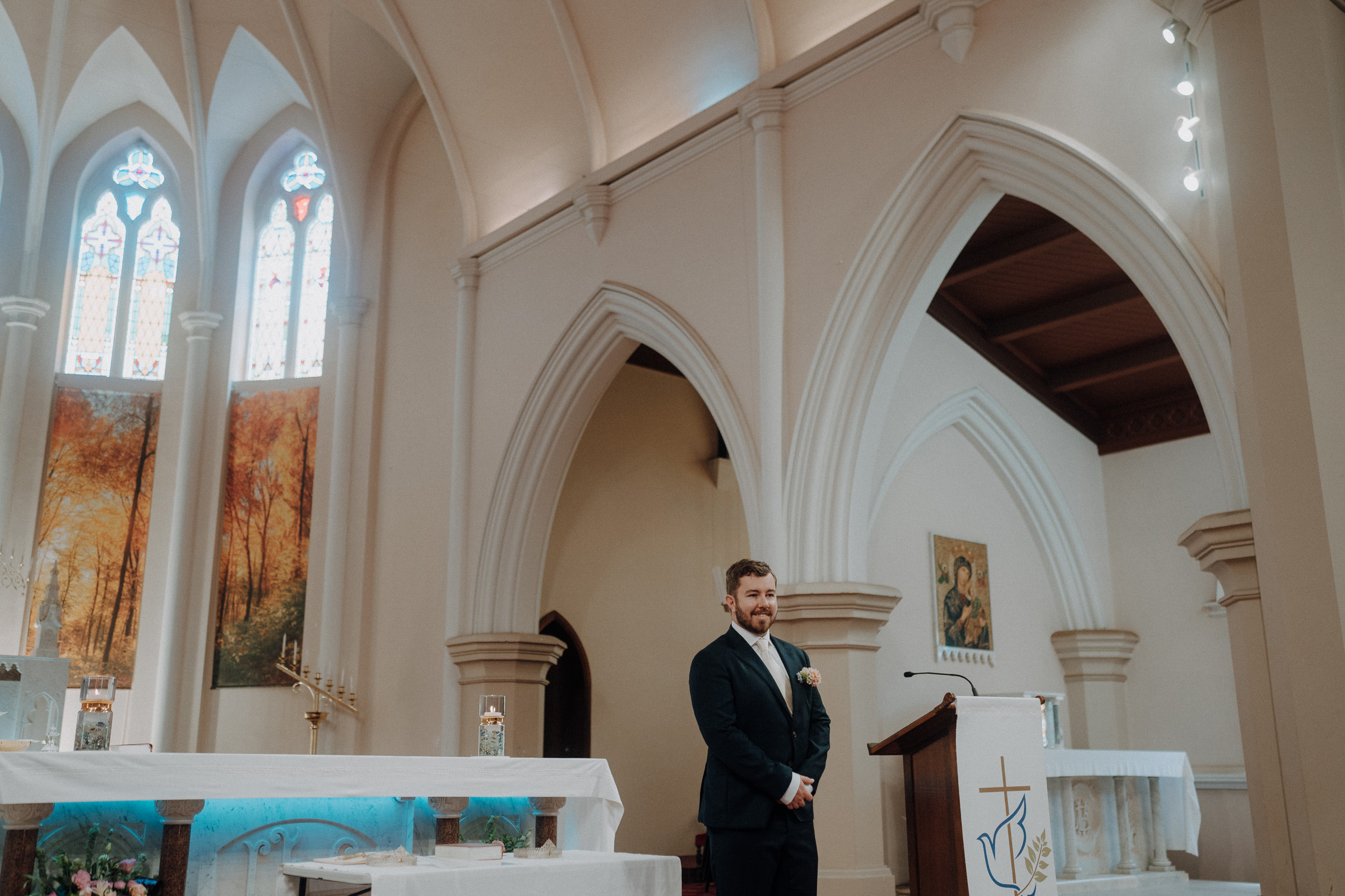 A person in formal attire stands near a podium in a church with arched ceilings and stained-glass windows.