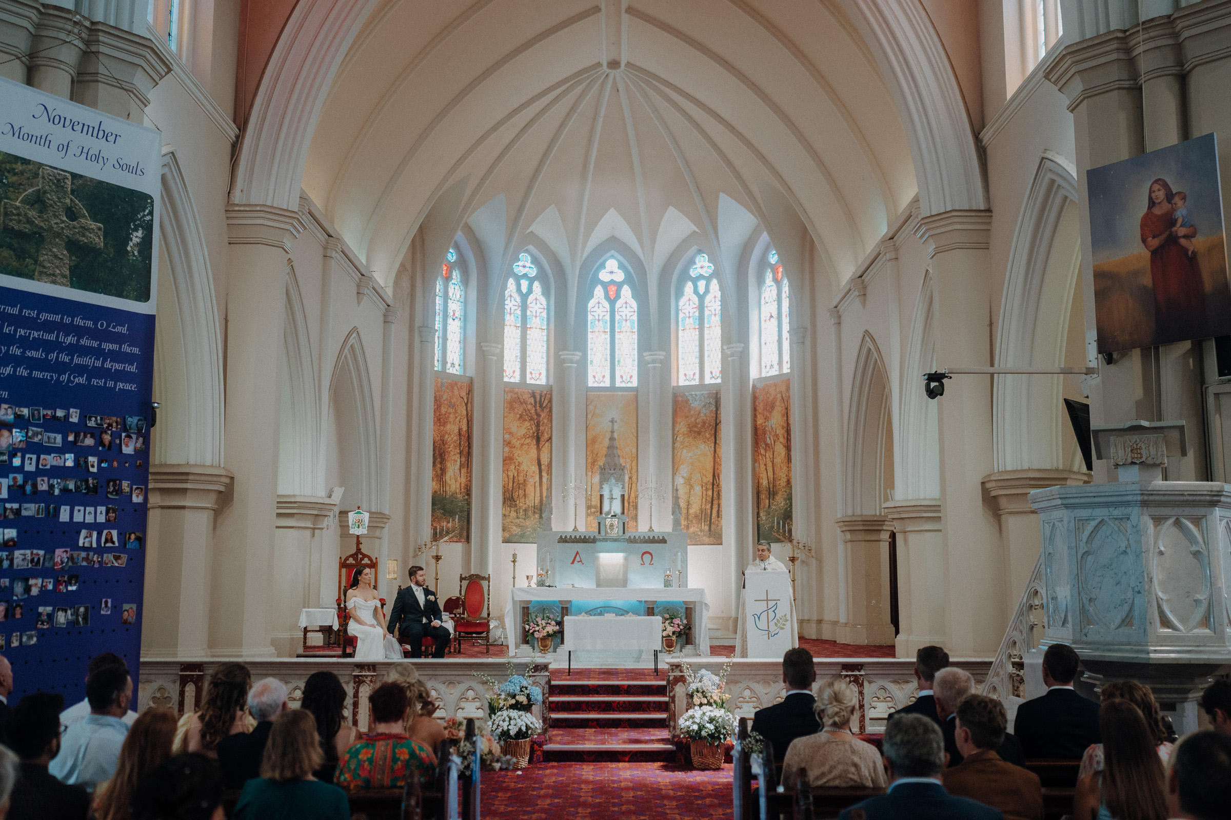 Interior of a church during a wedding ceremony. The bride and groom are seated at the altar with guests in attendance. Stained glass windows and religious decor are visible.